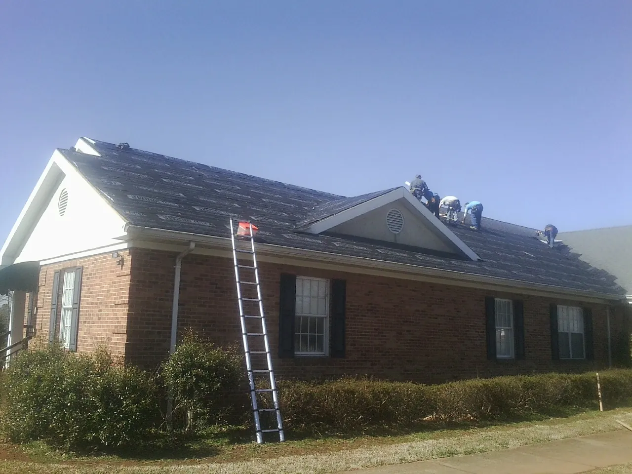 Crew tearing shingles off a brick ranch roof with a ladder set — Metal Roof Repair work by our crew in Wilmont