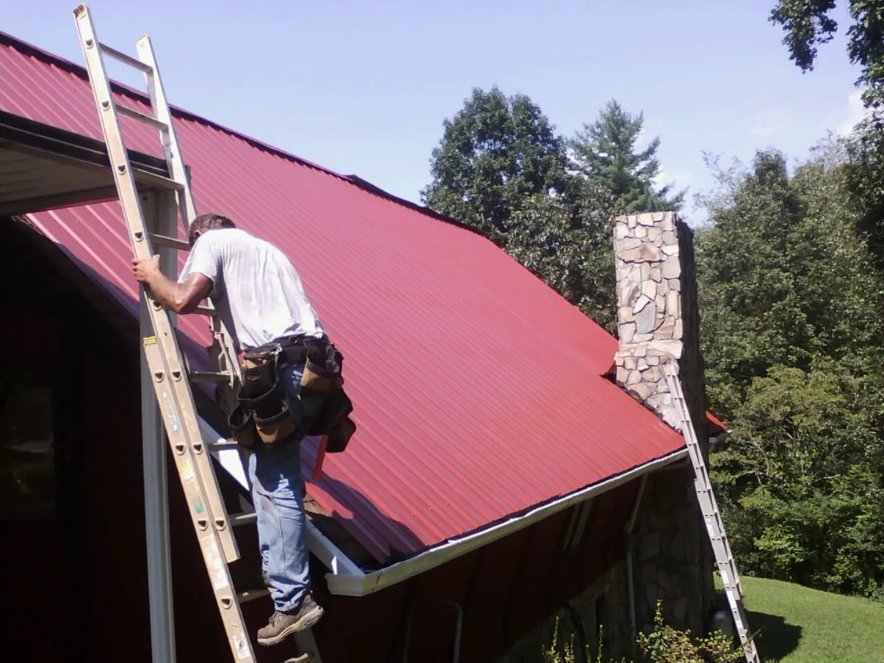 Roofer on a ladder beside a red metal roof and stone chimney — Soffit & Fascia Repair by Roofing Brothers in Wilmont